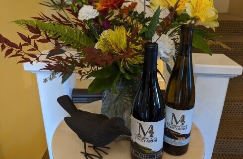 A small table with 2 bottles of wine, a bouquet of flowers, and a decorative black raven next to them.