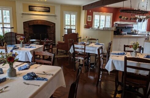 A dining room with several table set for either 2 or 4 people with white linen tablecloths, wood chairs, a fireplace along one wall, and a large opening into a kitchen