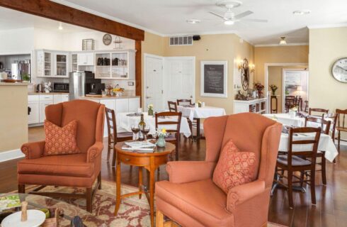 A dining room and sitting room area with several tables set for either 2 or 4 people, a kitchen off to the side, and a sitting area with 2 orange wing back chairs and a glass coffeetable