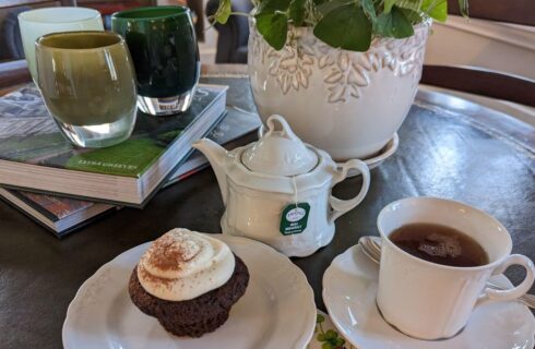 A table with a plate of a chocolate muffin, a teapot and a cup of tea, alongside a stack of books with candles on top.