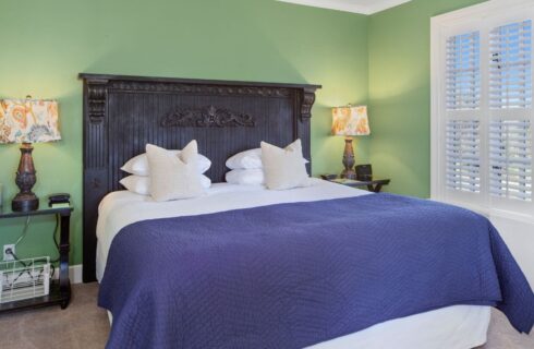 A bedroom with light green walls, white trim, beige carpet, a bed with a tall carved ornate wood headboard and white and blue bedding, and nightstands with lamps on either side of the bed, and a window letting in plenty of light.