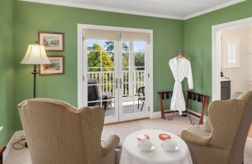 A bedroom sitting area with 2 wingback chairs, a round table in between them with 2 coffee cups, 2 bathrobes in one corner, a lamp in another corner, doors leading to a bathroom, and French doors leading to an outside balcony with white railings and green trees.