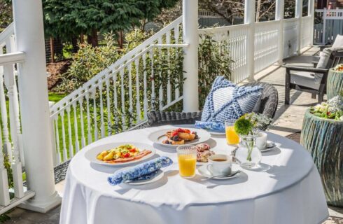 An outdoor porch with a table with white tablecloth set for breakfast with plates of eggs, bacon, french toast, and cups of coffee and glasses of orange juice.