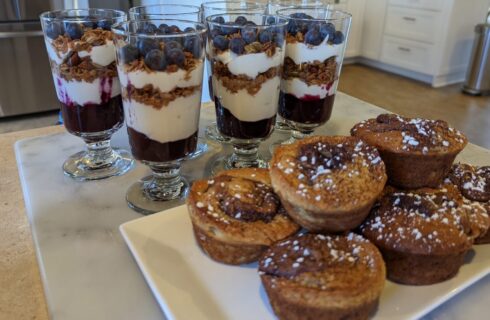 A table with a white plate of stacked muffins, and all parfait glasses with yogurt, granola, and fruit in them.