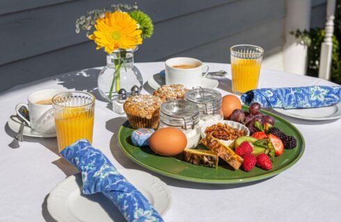 A table with a white tablecloth set for breakfast with a green plate of yogurt, granola, fresh fruit, muffins, cheese, and 2 hard boiled eggs, along with glasses of orange juice, cups of coffee, and a vase with flowers.