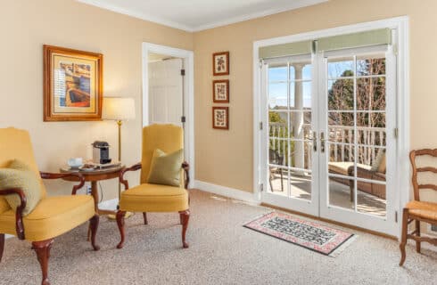 A bedroom sitting area with yellow chairs, a small table in between with an instant coffeemaker, coffee cups and plate of cookies, and French doors opening to out outside porch with lounge chair.