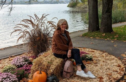 a woman sitting on a hay bale with pumpkins next to the hay, pink flowers nearby, and a lake in the background, with trees, bushes and green grass.