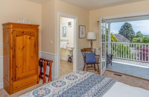 A bedroom with a bed with blue and white bedding, a wood wardrobe, a wood chair and lamp in the corner, a door open to a bathroom, and French doors opening out to a porch with white railing, green and red trees and the ocean in the background.