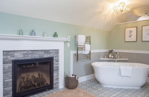 a bedroom with a deep soaking tub in the corner with towels on a rack, and a fireplace near it with a stone surround and a white wood mantle