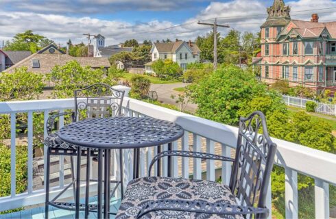 A balcony with white wood railings, a wrought iron table and 2 chairs, and multi story houses in the background with lush green trees and blue skies with white and grey clouds