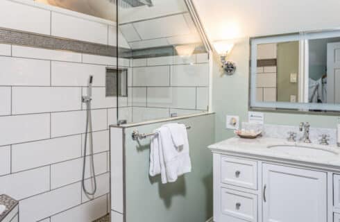 A bathroom with light green walls, a tiled shower, and a wood and granite vanity with a single sink, and long mirror above the sink.