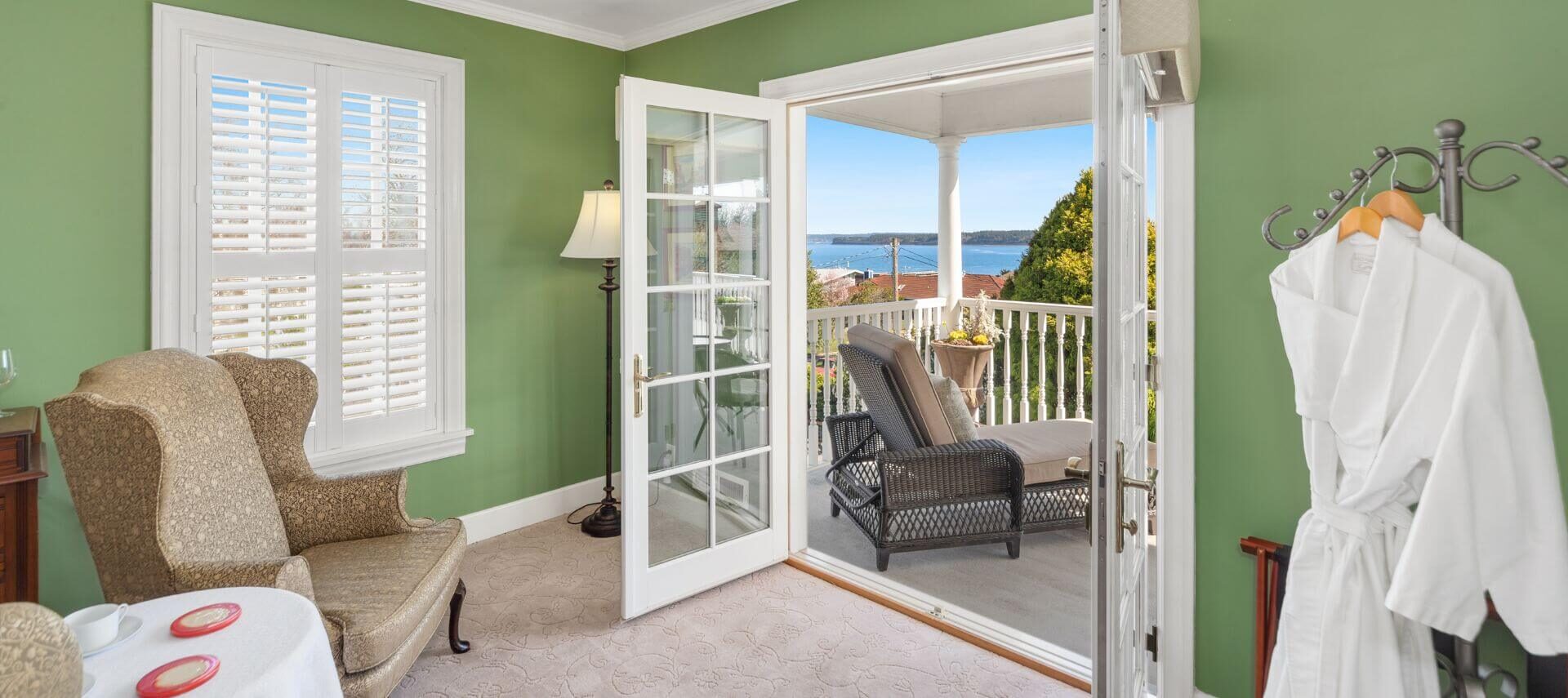 A sitting room in a bedroom with green walls and white trim, two wingback chairs on either side of a table with coffee cups, and French doors opening out to a balcony with lounge chairs and views of trees and the ocean