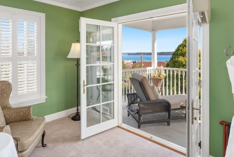 A sitting room in a bedroom with green walls and white trim, two wingback chairs on either side of a table with coffee cups, and French doors opening out to a balcony with lounge chairs and views of trees and the ocean