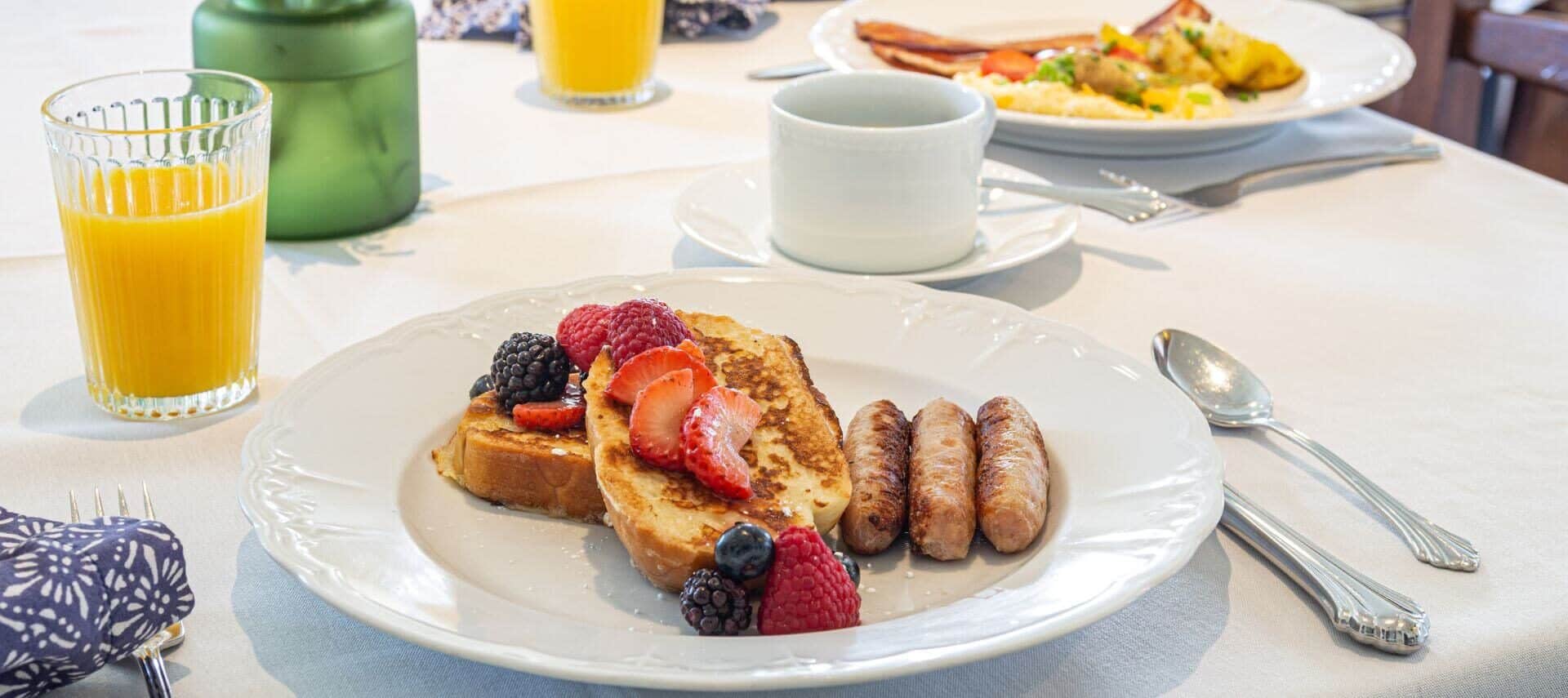 A table with a white tablecloth set for breakfast with plates of french toast topped with fruit, sausage, eggs, and bacon, along with a cup of coffee and glasses of orange juice.