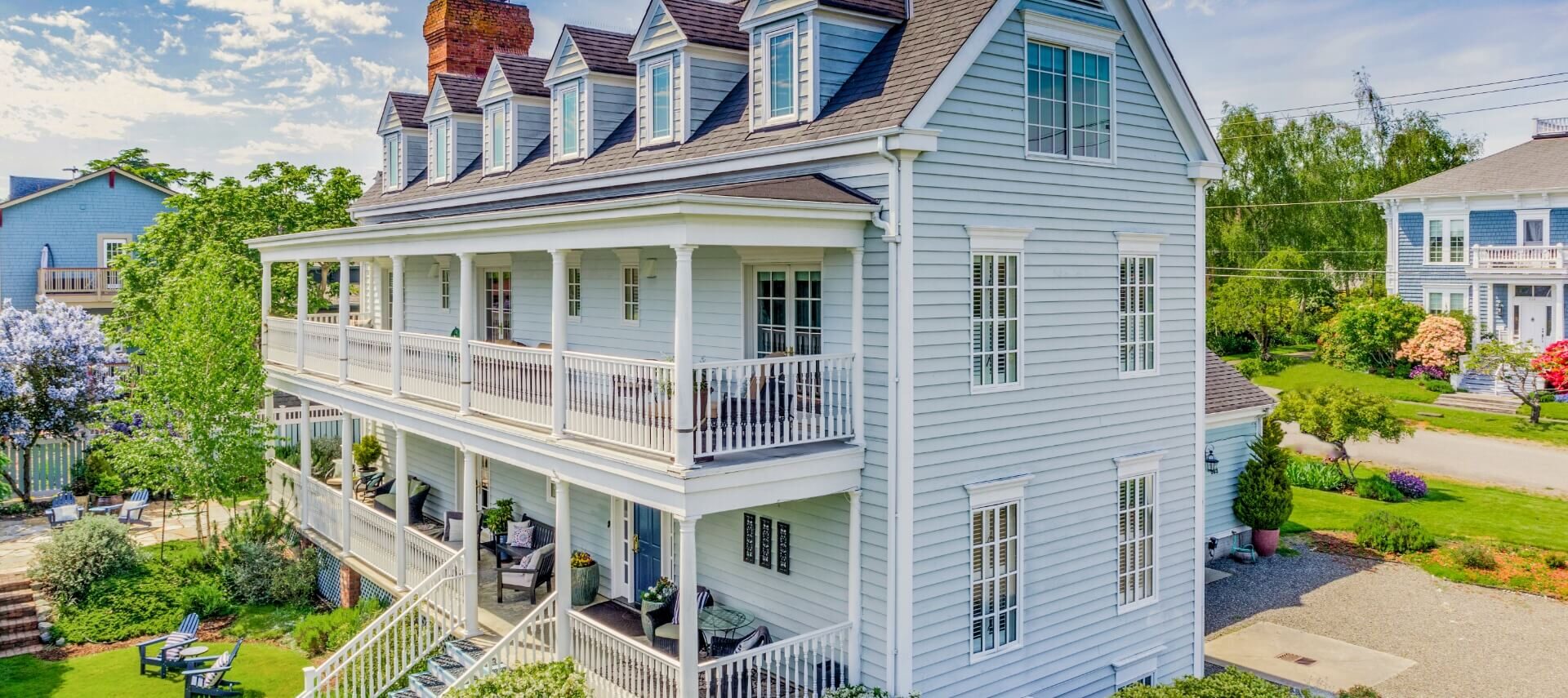 A multi story light blue house with porches, white trim, green grass, blue chairs and other houses in the neighborhood