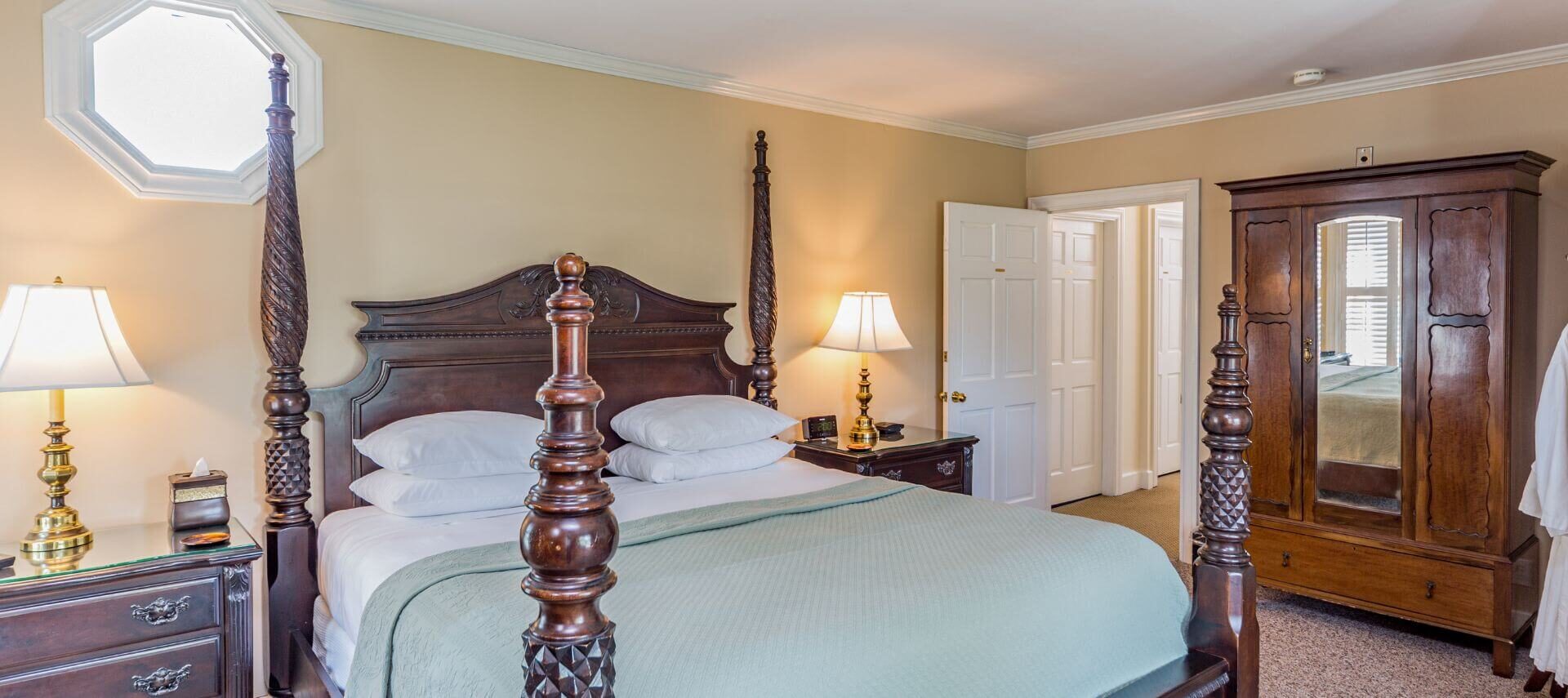 A bedroom with yellow walls with white trim, an ornate four poster bed with carved wood headboard, nightstands with lamps on either side of the bed, and aa wardrobe with mirror on another wall