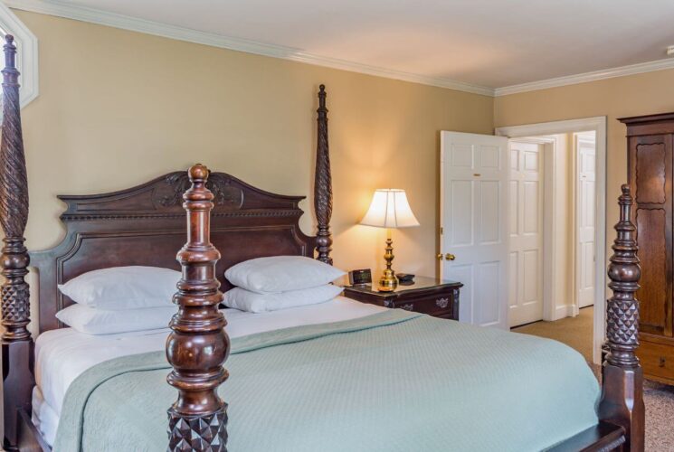 A bedroom with yellow walls with white trim, an ornate four poster bed with carved wood headboard, nightstands with lamps on either side of the bed, and aa wardrobe with mirror on another wall