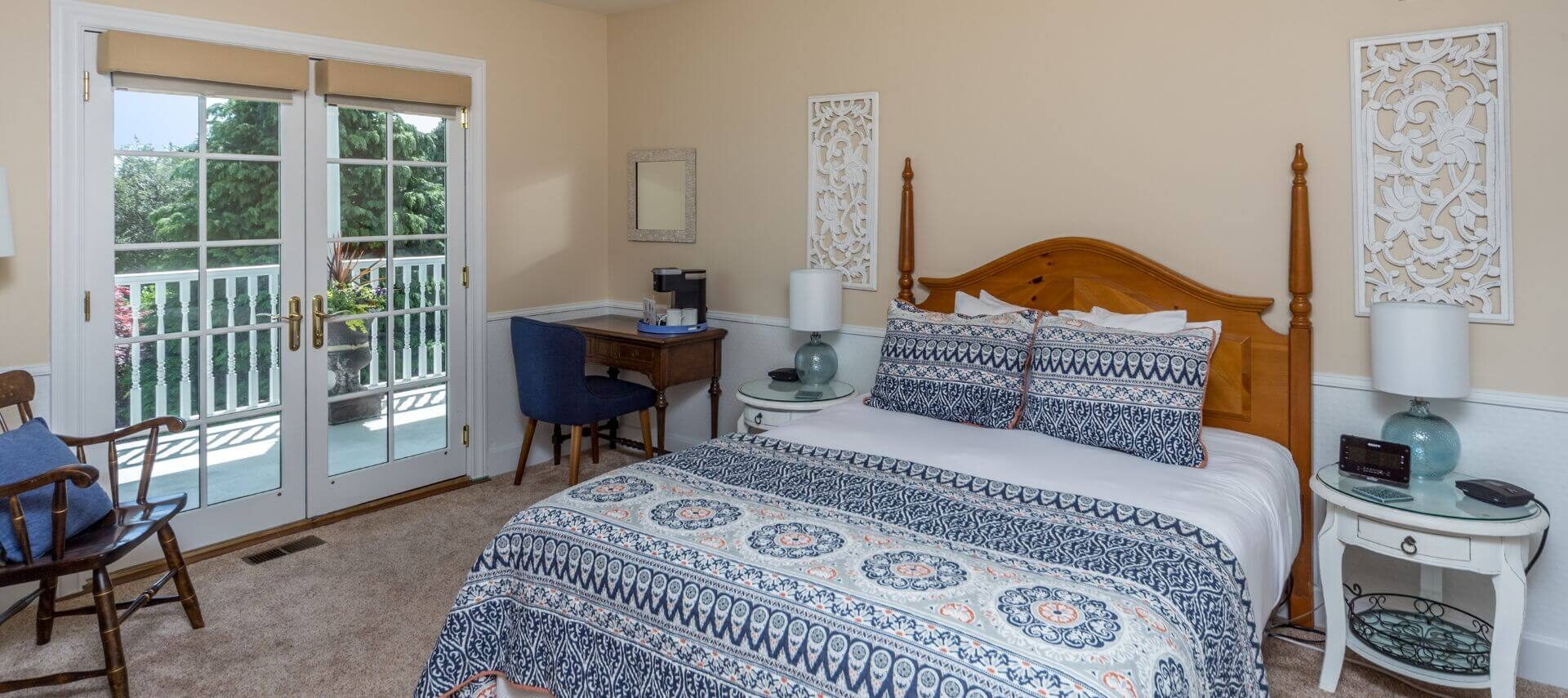 A bedroom with soft beige walls, white chair railing and trim, a wood bed with blue and white bedding, white wood nightstands with lamps on either side, and a wood desk and chair in the corner, along with French doors leading out to a porch with white railing and green treesh in the background.