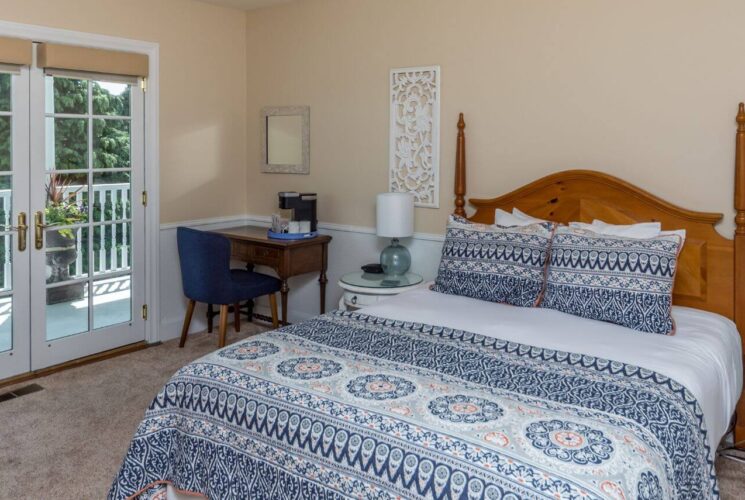 A bedroom with soft beige walls, white chair railing and trim, a wood bed with blue and white bedding, white wood nightstands with lamps on either side, and a wood desk and chair in the corner, along with French doors leading out to a porch with white railing and green treesh in the background.