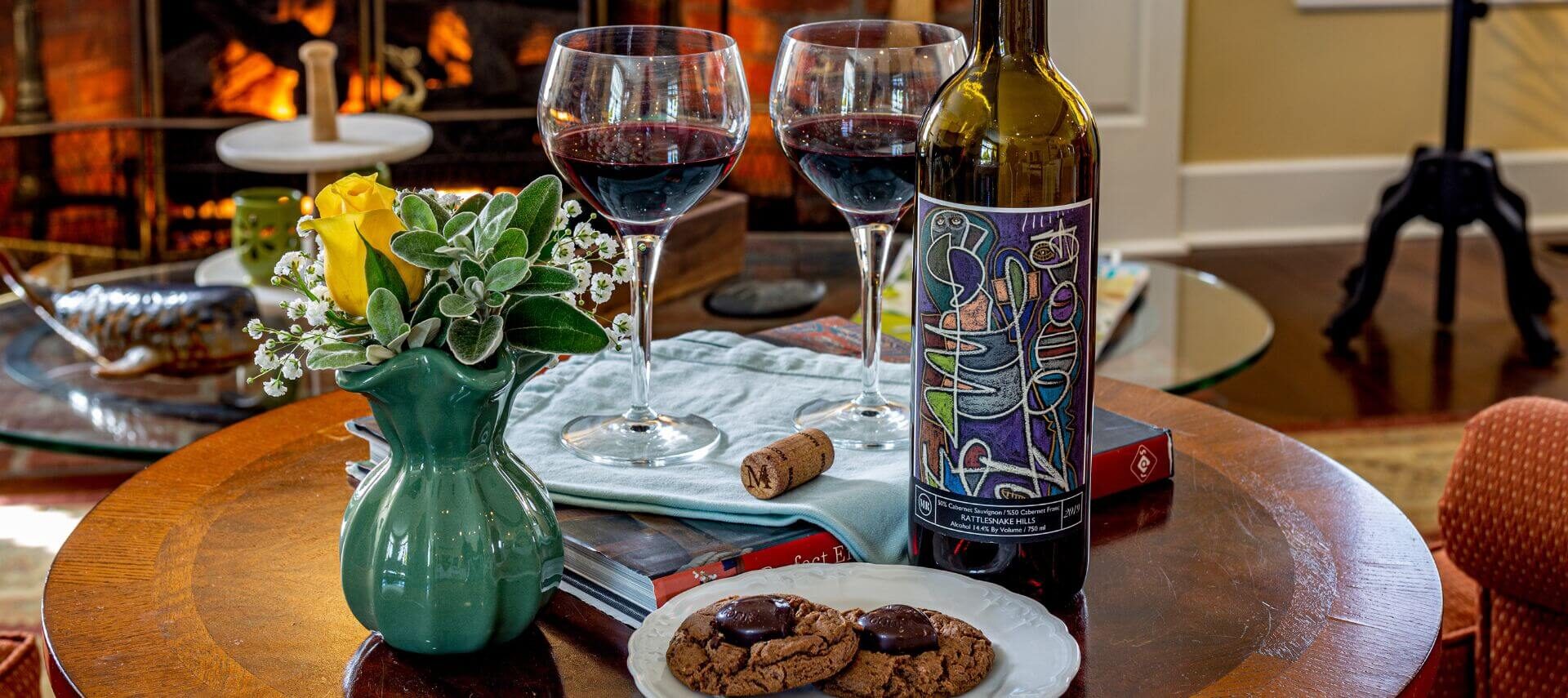 A table with a plate of chocolate cookies, a bottle of wine with 2 wine glasses filled with red wine, a green vase with a yellow rose, and a fireplace in the background.