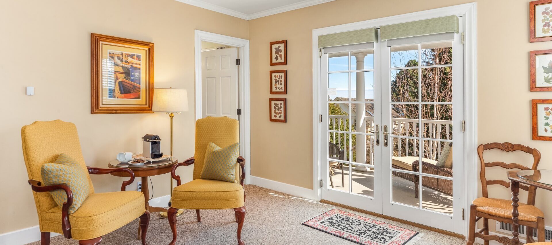 a sitting room with 2 yellow chairs, a side table in between them with a plate of chocolate cookies and a single serve coffee maker, and French doors opening out to a porch with patio furniture and trees in the background.