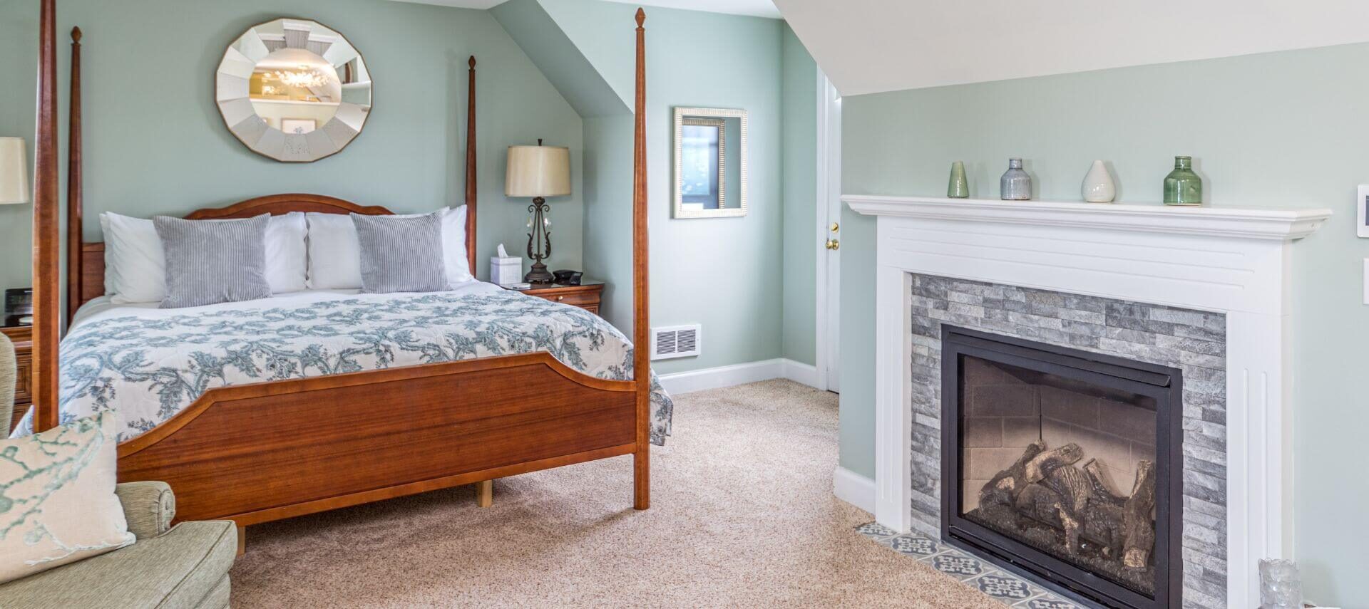 A bedroom with light blue walls, white trim, a large wood 4 poster bed with blue and white bedding, beige carpet, and a stone fireplace with a white wood mantle.
