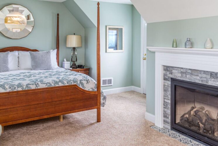 A bedroom with light blue walls, white trim, a large wood 4 poster bed with blue and white bedding, beige carpet, and a stone fireplace with a white wood mantle.