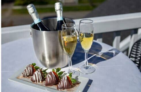 A table with a bucket of 2 splits of champagne, 2 champagne flutes with champagne in them, and a plate with 4 chocolate covered strawberries.