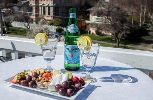 An outdoor white table with a platter of fruits and nuts, a bottle of Pellegrino sparkling water and 2 water glasses.
