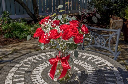 A vase full of red roses, greenery, white baby's breath, and a red ribbon, on a stone table with wrought iron chair in a garden.