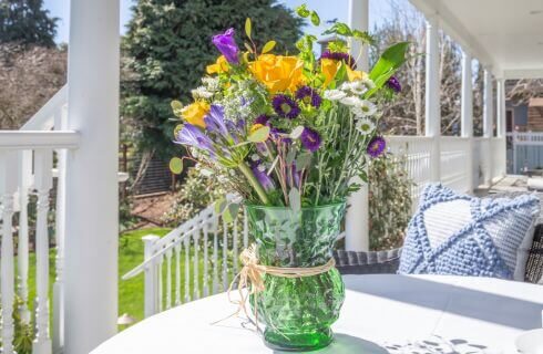 A green vase full of colorful wildflowers on a white table on an outside porch with white columns and railings.