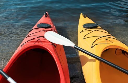 A red and yellow kayak with paddles on the shore of a lake.