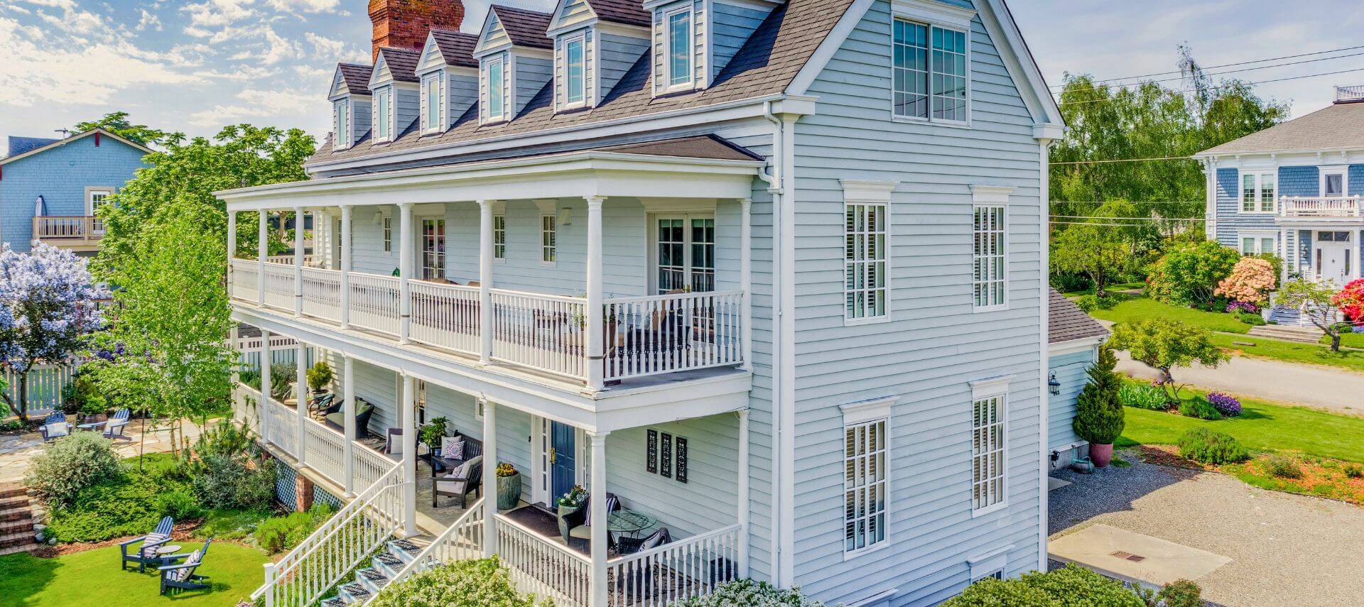 A multi story light blue house with porches, white trim, green grass, blue chairs and other houses in the neighborhood