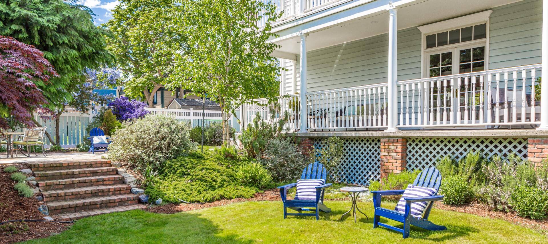 A light blue house with white trim, an expansive front porch, a green lawn with blue adirondack chairs, a stone patio with patio furniture and lush green trees and bushes.