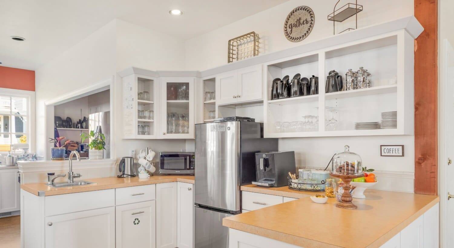 A country kitchen with white cupboards, laminate countertops, a stainless steel appliances, and plates, glassware, tea kettles and coffee pots on the shelves.