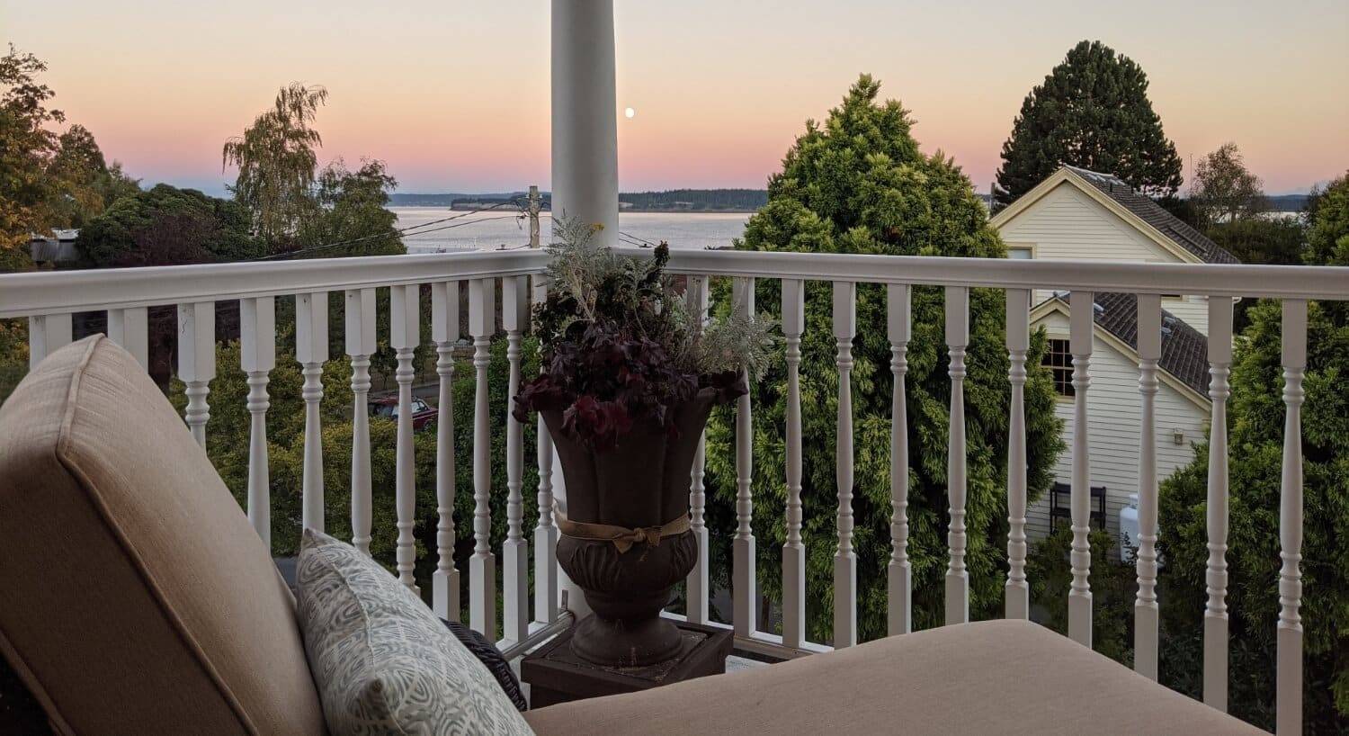 A lounge chair on a second story porch with white railings, green trees and another house, and the ocean in the background.