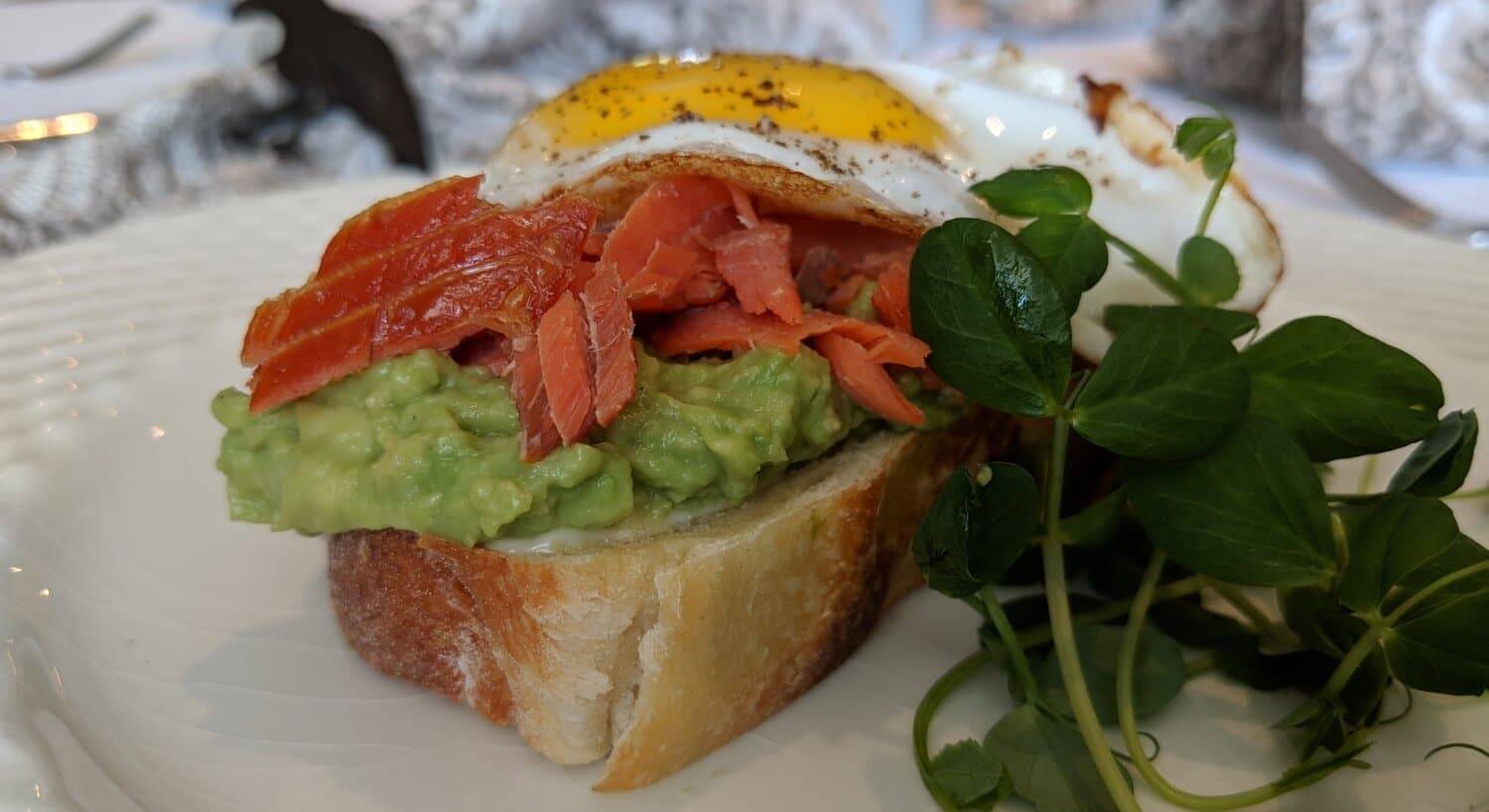 A thick piece of bread covered with mashed avocado, slices of smoked salmon, and topped with a sunny side ug egg, garnished with fresh herbs on the side.