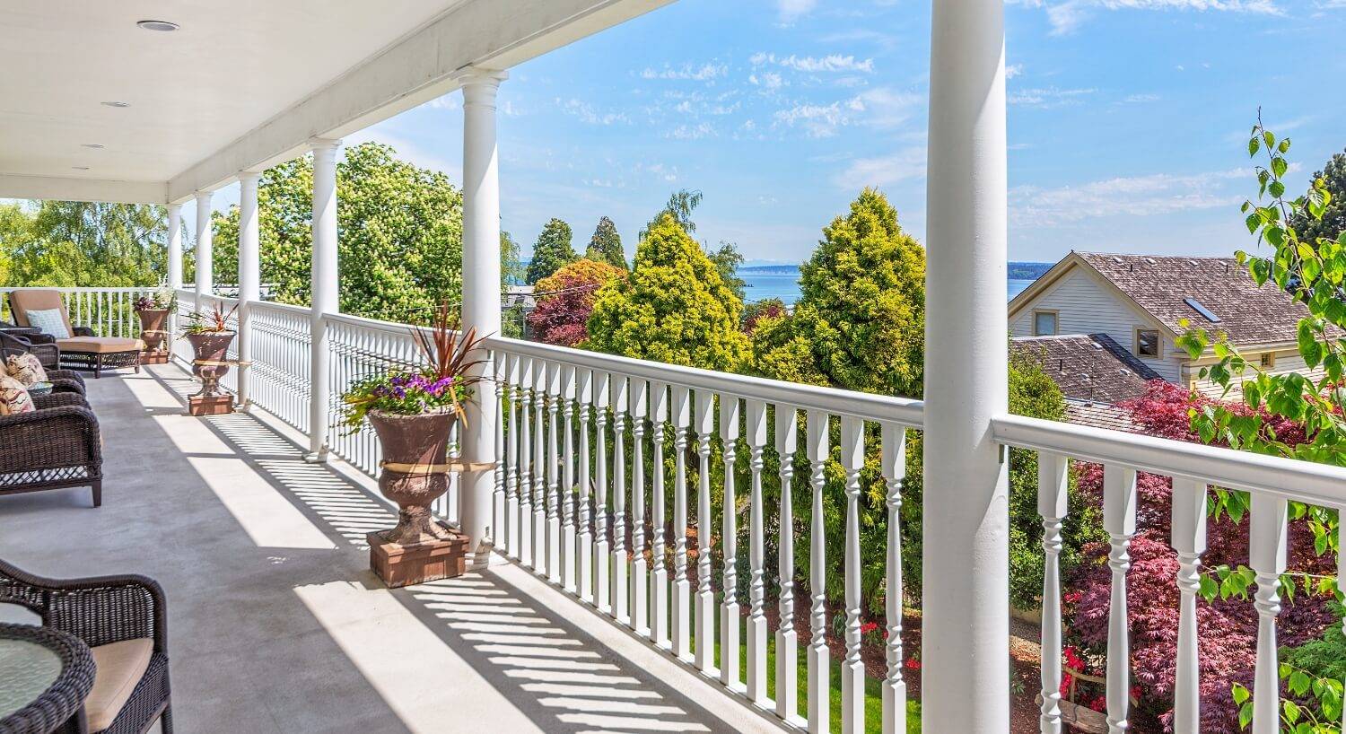 A sweeping second floor porch with wicker furniture, several planters with flowers, white railings and columns, trees, a house, and the ocean in the background.