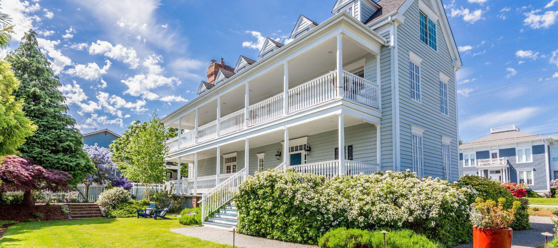 A multi story light blue house with porches, white trim, green grass, blue chairs and another houses in the backgroiund