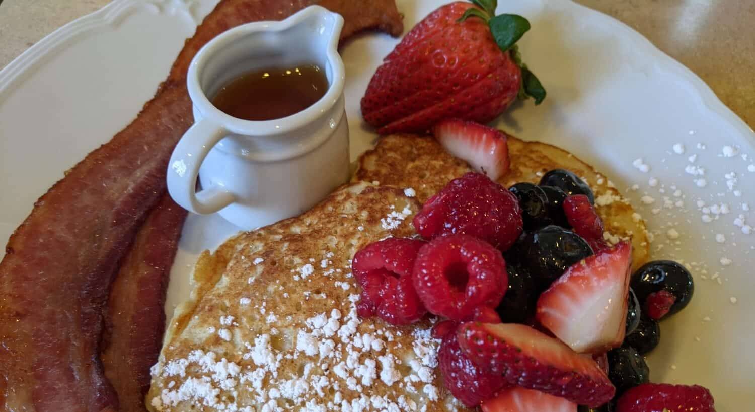 A plate with pancakes topped with fresh berries, 2 pieces of bacon, and a small pitcher of maple syrup.
