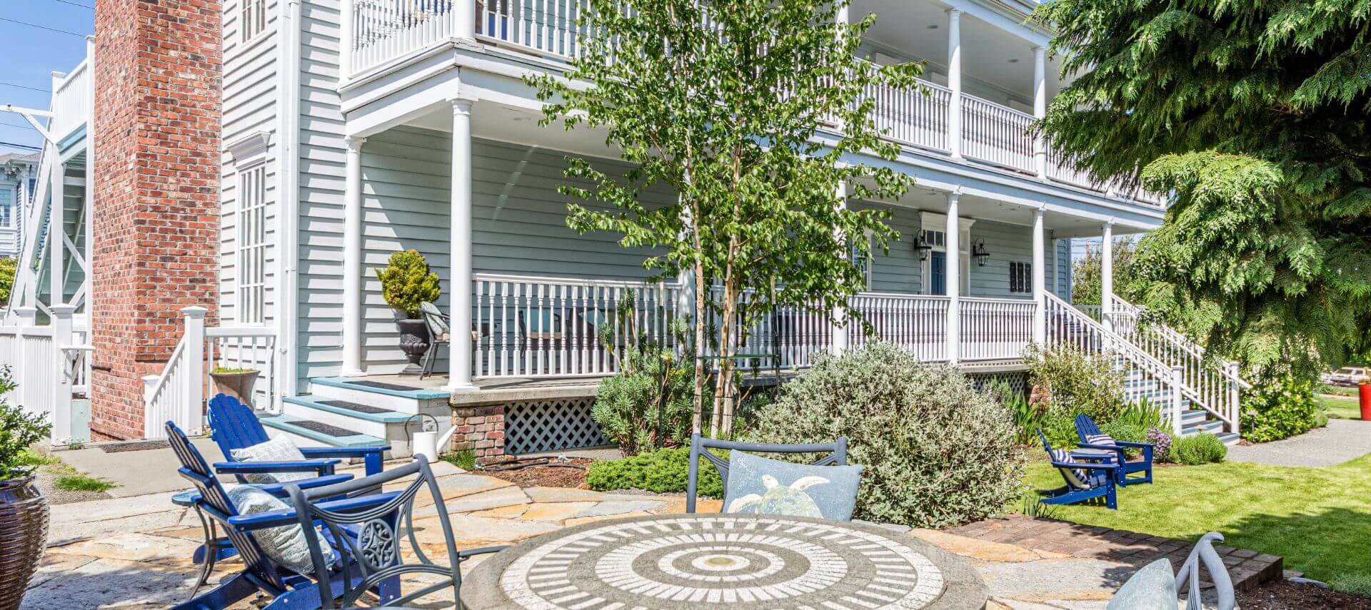 The front porches and sides of a multi story light blue house with white trim, green grass, a flagstone patio, a stone table and wrought iron chairs, and blue adirondack chairs