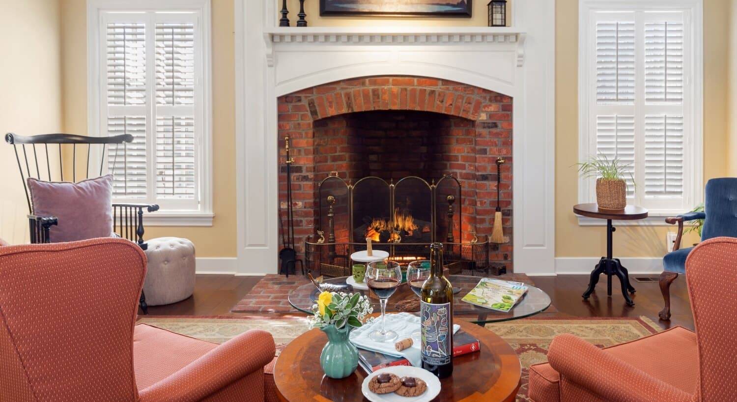 A sitting area with orange wing back chairs, a table in between then with a plate of cookies, a bottle of wine and 2 glasses of red wine, a vase of slowers, and a brick fireplace with a roaring fire along the wall.